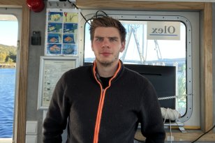 En mann med svart jakke står i en båt, han har mørkt hår og ser rett inn i kamera