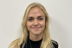 Lyshåret dame med svart t-skjorte og hvit jakke over smiler til kamera med en hvit vegg i bakgrunnen