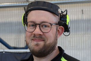 Ung mann med helskjegg og briller smiler til fotografen. På hodet har han hørselvern og han er ikledd en svart genser med gule striper på skuldrene. 