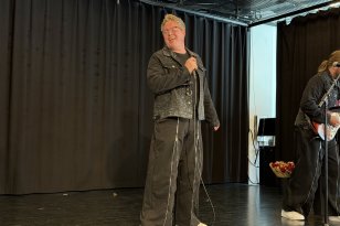 Mann med kort, blondt hår, iført svart bukse og svart jeansjakke, står på en scene og synger mens han holder en mikrofon. Bakgrunnen er mørk.