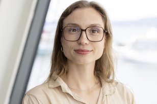 En dame med beige bluse og brunt hår smiler med munnen lukket. Hun har på seg briller. Bak henne er det en lys bakgrunn.
