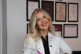 Kvinne med langt blondt hår iført hvit medisinsk frakk holder en svart notatblokk og en penn. På veggen bak henne henger flere innrammede diplomer.