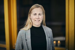 Advokat Marit Opsahl Jukvam En kvinne med langt blondt hår står ute foran et bygg med gule karmer. Hun smiler rett mot kamera. Hun har på seg en rutete blazer over en mørk genser.