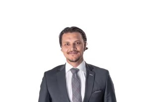 En mann med mørkt, bakovergredd hår og kort skjegg står foran en lys grå bakgrunn. Han har på seg mørk dress, hvit skjorte og slips, og ser rett inn i kamera med et rolig smil.