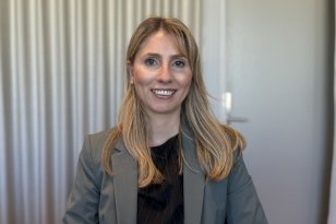 En kvinne sitter vendt mot kamera i et kontormiljø og smiler vennlig. Hun har langt, lyst hår som faller over skuldrene og er kledd i en mørk topp under en grå dressjakke. Bak henne henger lyse gardiner som dekker en vegg eller et vindu, og en dør er synlig i bakgrunnen.