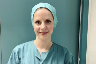 Portrettbilde av en dame i grønn operasjonsklær. Hun står foran en grønn dør. I arbeidsskjorten har hun et skilt som sier hun er operasjonssykepleier 