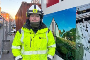 En anleggsrørlegger står ute på en byggeplass, kledd i gul refleksjakke og hjelm med hodelykt. Han ser rett mot kamera og står ved siden av en arbeidsbil med firmalogo og bilder av ferdige prosjekter.
