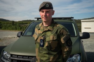 En person i militæruniform med beret står foran et militært kjøretøy og ser mot kameraet. Bakgrunnen viser åpent landskap og en parkeringsplass.
