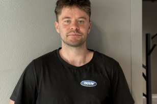 En mann står foran en grå vegg og ser mot kameraet. Han har på seg en svart T-skjorte med Hasle-logo på brystet.