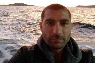 En mann med mørkt kort hår tar en selfie på en båt i solnedgangen. Bak ham er sjøen og to lave øyer i horisonten, mens himmelen er lys med noen mørke skyer.