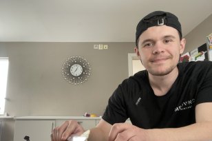 En ung mann i svart t-skjorte og svart caps sitter ved en kjøkkenbenk og smører pålegg på en brødskive