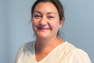 Kvinne i hvit bluse står innendørs og smiler til kamera foran en gråblå bakgrunn