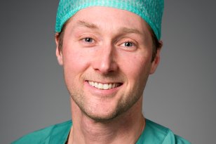 En mann i grønt operasjonstøy og operasjonshette smiler mot kameraet i et portrettbilde. Han har lys hud, kort hår som er delvis skjult under hetten og står foran en nøytral grå bakgrunn. 