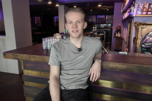 En blond mann med grå t-skjorte sitter lett henslengt foran en bardisk. Bak baren står det diverse flasker og glass
