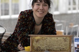 Dame med mørkt hår og mørk overdel står foran en kasse i isopor og en honningtavle. I bakgrunnen er det store glassvinduer.