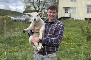 En mann i rutete skjorte og arbeidsbukse står ute på et jorde og holder et lam i armene. Han smiler mot kamera. I bakgrunnen er det gress, trær, biler og et lyst gårdshus under en skyet himmel.
