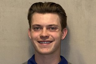 En ung mann med grå genser og mørk blondt hår står foran en lys vegg.