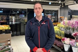 En mann med kort hår og mørk hettegenser står foran et blomstertorg med rosa og gule blomster. 