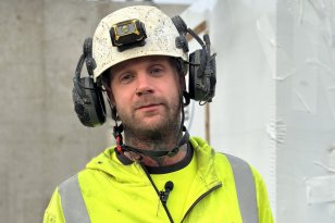 En byggarbeider med litt skjegg står utendørs på en byggeplass. Han har på seg en gul refleksjakke med logoen «Con Form» på brystet, og en hvit hjelm med hodelykt og hørselsvern. Han ser rett mot kameraet og smiler svakt. Bak ham sees en grå betongvegg og deler av en hvit innpakning.