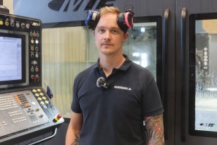 En person står foran en stor CNC-maskin med kontrollpanel. Han ser inn i kameraet og har hørselsvern på hodet. CNC-maskinen bak dem har store vinduer og kontrollskjerm med knapper og måleindikatorer. Personen har på mørkeblå arbeidsklær og tatoveringer på armen.