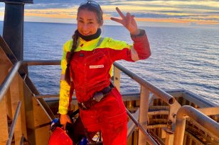 En ung kvinne i rødt offshorearbeidstøy står på en plattform med havet og en dramatisk solnedgang i bakgrunnen. Hun smiler mot kameraet og holder opp et fredstegn med hånden, mens hun holder vernehjelmen i den andre.