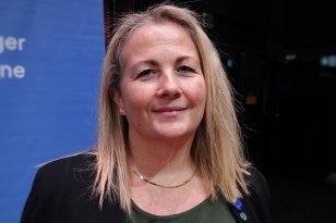 Kvinne med lyst hår i svart dressjakke, grønn bluse og gullsmykke smiler rett frem til kamera med blå bakgrunn.