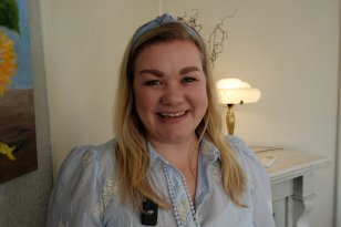 Kvinne med blondt hår og blå bluse med hårbølje. Hun smiler til kamera med et bilde på veggen ved siden av.