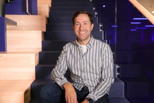 En mann sitter avslappet på en mørk trapp innendørs, med et varmt smil mot kameraet. Han har på seg en lys, stripete skjorte og mørke olabukser. Bak ham skimtes moderne kontorinteriør med blått lys og glassrekkverk.