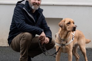 En mann i mørke klær. Han smiler og ser rett i kamera. Han sitter på huk og holder en førerhund i bånd. Det er en lys labrador. De er ute.
