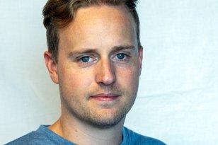 En mann med blondt hår og blå genser står foran en lys vegg.