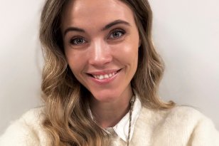 Dame med blondt hår og hvit genser står foran en hvit vegg og smiler til kamera