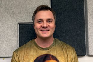 En mann med kort mørkt hår står vendt rett mot kameraet og smiler svakt. Bak ham er grå og mørke akustiske lydpaneler montert på veggen.