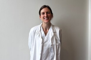 En kvinne med mørkt hår står i hvit frakk foran en hvit vegg