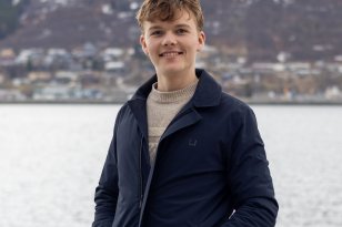 En mann i lys genser og mørkeblå jakke. Han ser rett i kameraet og smiler. Han er ute, bak han er havet, hus og fjell.