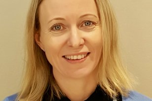 Portrettbilde av en kvinne som ser rett mot kamera og smiler. Hun har skulderlangt blondt hår og har på seg lyseblå arbeidsklær med mørk krage og navneskilt. Bakgrunnen er lys og ensfarget.