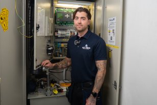 En mann står foran et elektrisk skap og smiler til kamera. Han har brunt hår og mange tatoveringer på armene. Han har på en mørkeblå t skjorte der det står "ALT heis"