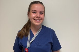 En ung kvinne med brunt hår i hestehale og blå skjorte står foran en lys vegg.