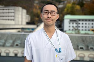 En mann i hvit legeuniform med mørkt kort hår, som står utendørs foran sykehusbygninger med fjell i bakgrunnen. Han ser rolig og profesjonell ut, med stetoskop i lommen og ID-bånd rundt halsen.