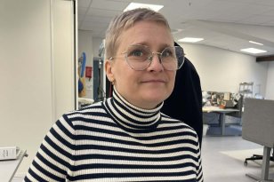 Korthåret dame med briller og stripete genser ser inn i kamera. Hun står i et hvitt rom med pulter og symaskiner i bakgrunnen.