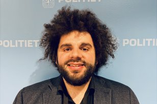 Mann med langt, mørkt og krøllete hår og langt skjegg står og smiler i et portrettbilde. Han har på seg svart skjorte og mørkegrå dressjakke. I bakgrunnen sees en lyseblå vegg med politilogo.