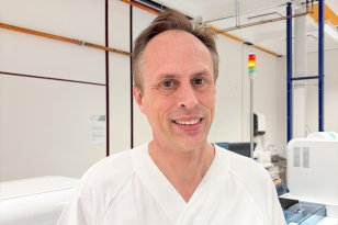 Mann med kort, mørkt hår i hvit sykehusuniform smiler mot kamera i et portrett. I bakgrunnen står ulike laboratoriemaskiner og utstyr.