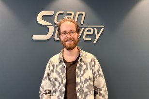 Mann med oransjbrunt hår, skjegg og briller står og smiler mot kamera. Han har på en svart T-skjorte og en åpen, hvit-grå mønstret genser. I bakgrunnen vises en vegg med logoen til Scan Survey.