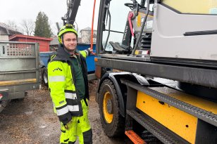 Anleggsmaskinfører i neon arbeidsklær og hjelm står ved en gravemaskin og poserer.