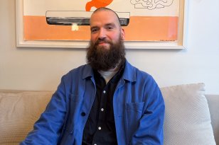 Mann med kort hår og langt mørkt skjegg, iført lyseblå jakke og mørkeblå skjorte, sitter i en sofa og poserer. I bakgrunnen henger en illustrasjon på veggen.