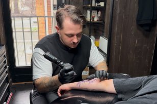 Mann med mørkt hår, hvit T-skjorte og svart vest holder en tatovørmaskin og gjør seg klar til å tatovere en kunde som ligger med armen frem. I bakgrunnen sees tatovørstudioet.