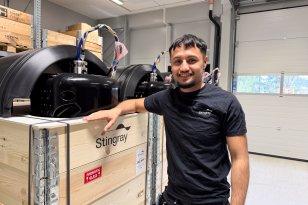 Mann med kort svart hår, svart arbeids T-skjorte og vernebukse står ved ferdigstilte produkter som er klargjort for utsending til kunder.