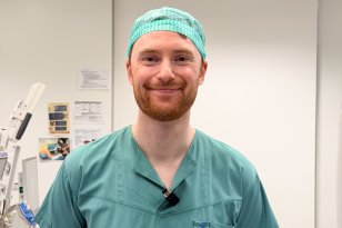 Mann med oransje skjegg iført grønt kirurgisk arbeidstøy og kirurgisk hette står og smiler mot kamera.