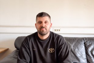 Mann med kort svart hår, svart genser og svart arbeidsbukse sitter i en skinnsofa i et pauserom på arbeidsplassen og poserer til et bilde.