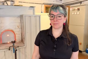 Kvinne med langt mørkt hår og turkis pannelugg, iført briller med klart glass, svart T-skjorte og svart bukse, står og poserer til portrett i et glassblåserverksted. I bakgrunnen sees utstyr og arbeidsmiljø fra verkstedet.