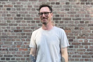 Her ser du kurator Pål Henrik. Han bruker mye tid på Preus Museum som museets mangfoldskurator. Mann med lys grå t-skjorte og briller står foran en murvegg og smiler.